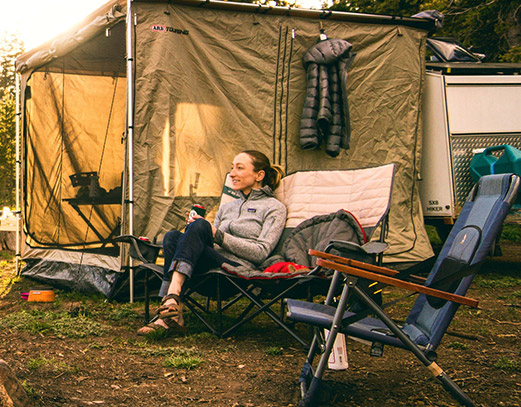 Woman smiling and relaxing in a camping chair, sitting next to another chair.